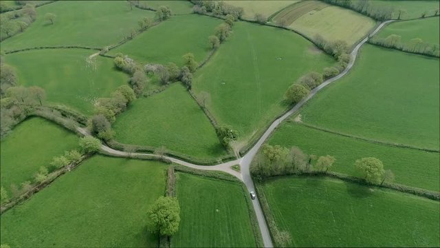 Aerial of a silver car travels down an English country road and passes a triangular junction. Green fields of spring and hedgerows surround the vehicle.