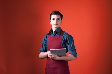 handsome barista in burgundy apron looking at camera and holding digital tablet on orange background with copy space