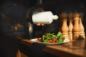 Cook pours olive oil on a fresh salad of tomato and green salad. Lighting the hand and the salad by the sunbeam from the window, the low key
