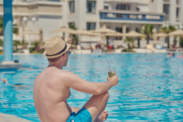 European man in a sun hat is relaxing in the pool of luxurious hotel. He is drinking cocktail and enjoying his holidays.