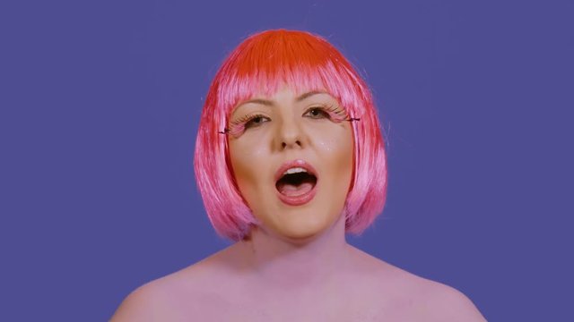 Stylish young woman back vocalist sings la la la and looks directly into the frame on a blue background. A girl with a bright make-up and red-pink hair performs vocals emotionally