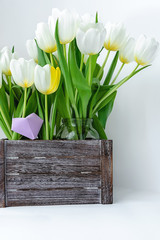 A bouquet of white tulips in a wooden box and a paper heart of lilac color on a white background.