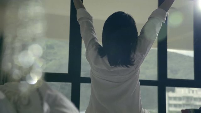 rear view of asian woman sitting on edge of bed stretching arms and body looking at view