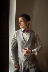 The fashionable bridegroom expects the bride near the window. Portrait of the groom in a grey vest
