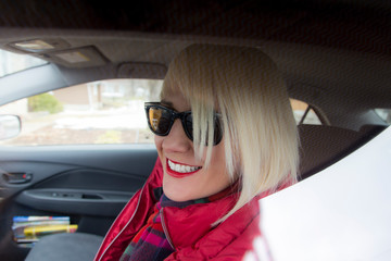 Horizontal closeup of pretty smiling platinum blonde young woman dressed in red and wearing sunglasses seen framed by closed car window