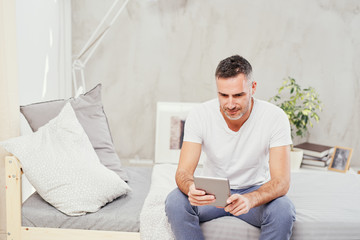 Caucasian man in forties sitting on bed in bedroom and using tablet.