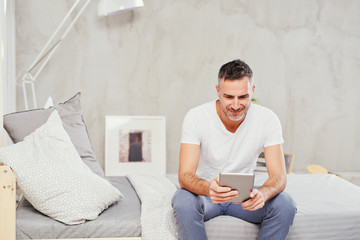 Caucasian man in forties sitting on bed in bedroom and using tablet.