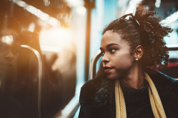 The portrait of a young charming African-American female pensively looking outside the carriage window while sitting indoors of a metro train; Brazilian girl in a subway train, shallow depth of field - Powered by Adobe