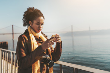 A pensive young biracial female tourist is leaning against an iron fence outdoors on the quayside while answering a message via the smartphone about her travel, with a bridge in the background