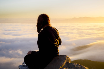 young woman sitting in the mountains over the clouds during sunrise