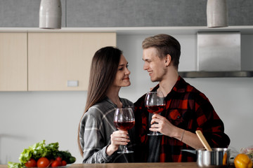 Beautiful young couple in plaid shirts in the kitchen preparing food and drinking red wine from glasses.