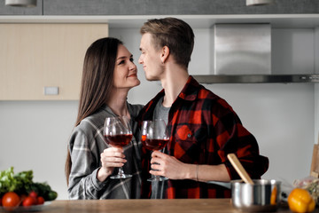Beautiful young couple in plaid shirts in the kitchen preparing food and drinking red wine from glasses.