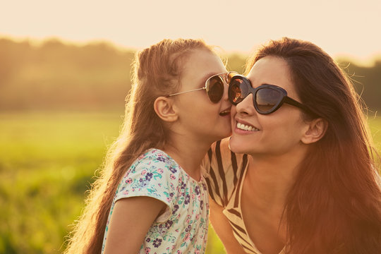 Happy fashion kid girl whisper the secret to her mother  in ear in trendy sunglasses in profile view on nature sunset background. Closeup