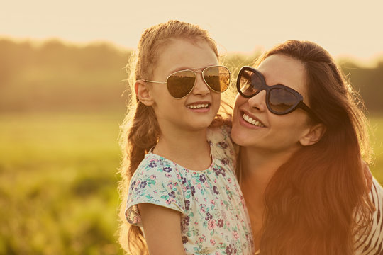 Happy fashion kid girl whisper the secret to her mother  in ear in trendy sunglasses in profile view on nature sunset background.
