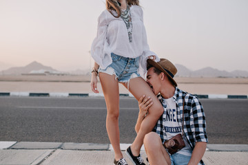 Young man in trendy shirt and vintage hat sitting on the road, funny kissing leg of his girlfriend. Slim girl in denim shorts fooling around with her boyfriend, spending time outdoor near highway - Powered by Adobe