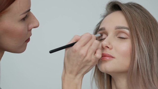 Makeup artist makes makeup beautiful blonde girl on a light monophonic background. Paints eyes with mascara, applies powder with a brush. Makeup artist studio concept.