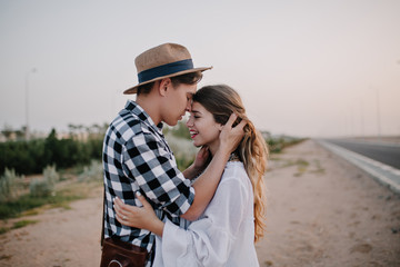 Fototapeta premium Excited cheerful girl in white tunic soft embracing boyfriend in stylish shirt, while he touching her long hair. Young couple of travelers hugging standing near the road and enjoys evening date