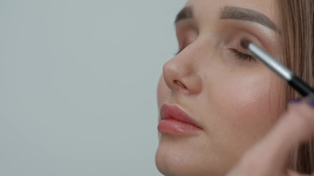 Makeup artist makes makeup beautiful blonde girl on a light monophonic background. Paints eyes with mascara, applies powder with a brush. Makeup artist studio concept.
