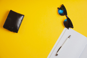 Top view of wallet, sunglasses and notebook with pen on yellow background