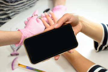 Blank mobile phone screen for text. A woman using a mobile phone, receiving a procedure for manicure and nail care by a beautician. A woman receives nails, manicure in a beauty salon.