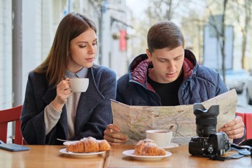 Young tourists man and woman reading map of city in outdoor cafe. Couple drinking coffee tea and eating croissants, spring city background