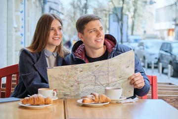 Young tourists man and woman reading map of city in outdoor cafe. Couple drinking coffee tea and eating croissants, spring city background