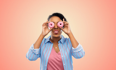 people, fast food and fun concept - happy african american young woman with donuts instead of eyes over living coral background