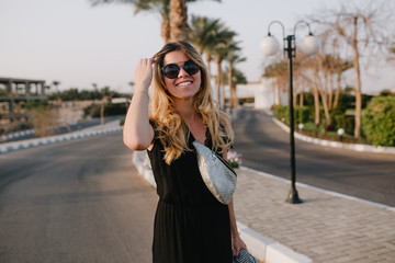 Attractive blonde woman in black dress and stylish sunglasses posing outside with exotic palm trees on background. Portrait of young woman with cute hairstyle and silver bag walking down the street