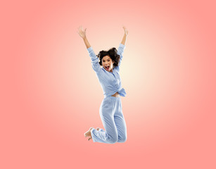 Fototapeta na wymiar fun, people and bedtime concept - happy young woman full of energy in blue pajama jumping over living coral pink background