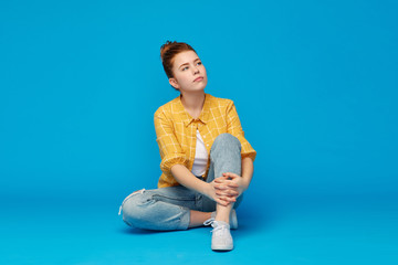 people concept - red haired teenage girl in checkered shirt and torn jeans sitting on floor over bright blue background