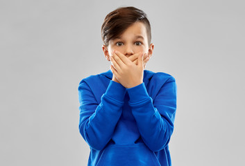 childhood, expressions and people concept - shocked little boy in blue hoodie closing his mouth by...