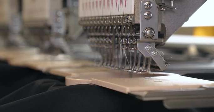 In slow motion video in a garment factory, a sewing machine embroiders various companies and engravings. Concept of: Automated work, Sewing machine, Engraving, Thread, Fabric, Needle.