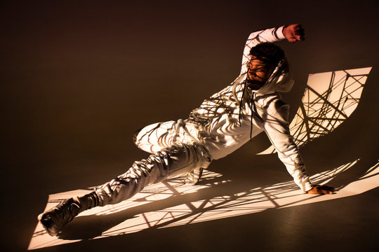 Handsome African American male in sportswear performing dance movement under bright light from window on brown background