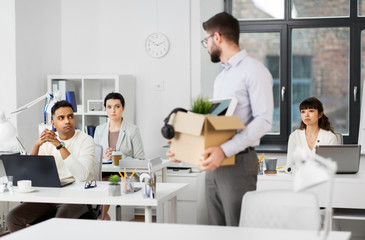 business, firing and job loss concept - fired male office worker with box of personal stuff looking at his sad colleagues