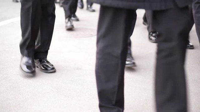 Many men in identical black patent leather shoes and black trousers walk down the street.