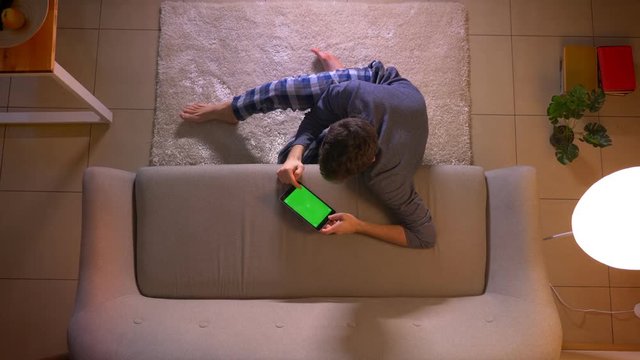 Closeup top shoot of young man watching an advertisement the tablet with green screen while sitting on the floor indoors at cozy home