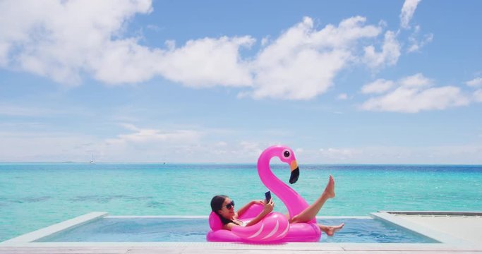 Vacation woman on inflatable Flamingo float mattress using mobile cell phone in swimming pool. Girl relaxing sunbathing enjoying travel holidays at resort pool in bikini. Luxury lifestyle.
