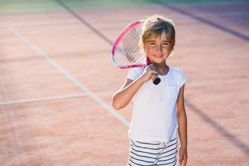 Happy little girl dressed white uniform with tennis racket on the shoulder on the background of outdoor tennis court.