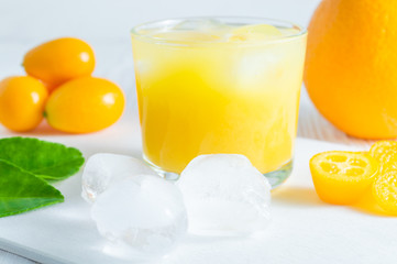 Refreshing summer citrus drink with orange, kumquat and ice cubes on white wooden table.