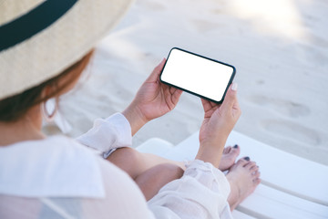 Mockup image of a woman holding white mobile phone with blank desktop screen while laying down on beach chair on the beach