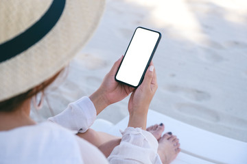 Mockup image of a woman holding white mobile phone with blank desktop screen while laying down on beach chair on the beach