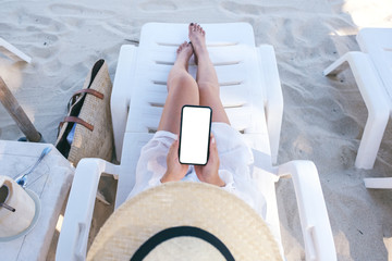 Top view mockup image of a woman holding white mobile phone with blank desktop screen while laying...