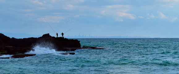Naklejka premium silhouettes de pécheurs sur un rocher au bord de la mer avec Beyrouth au loin, sur la ligne d'horizon