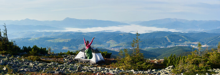 Panoramic view of camping on the top of mountain in morning. Back view of hiker woman in pink sweater and sleeping bag resting in tourist tent, holding hands lifting up. On foggy mountains background.