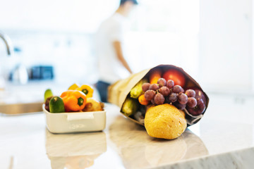 Colorful fresh vegetables and fruits with a bag of groceries shopping in the kitchen.Young handsome man cooking in background. Healthy eating concept.