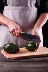 A young woman in an gray apron cuts an avocado on a wooden cutting board
