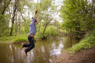 Cute boy rides a homemade bungee in the park. Bungee tied to a tree branch on the river bank. A...
