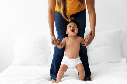 Laughing baby standing up on bed while holding his mother's hands