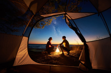 View from inside tourist tent. Young romantic couple, man and woman sitting at campfire holding hands on sea shore on evening starry sky at sunset and sea water background. Tourism and love concept.