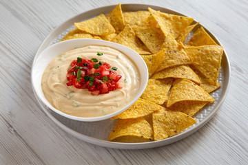 Homemade cheesy dip in a bowl, yellow tortilla chips over white wooden surface, side view. Close-up.